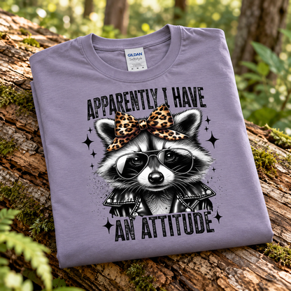 A folded paragon Gildan t-shirt with a graphic of a raccoon wearing sunglasses and a leopard-print bow tie, resting on a mossy log in a forest setting.