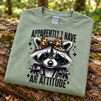 A folded sage green Gildan Softstyle t-shirt is displayed on a mossy log in a forest setting. The shirt features a graphic of a raccoon wearing sunglasses and a leopard-print bow, with the text 'APPARENTLY I HAVE AN ATTITUDE'.