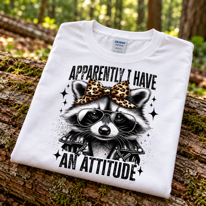 A folded white Gildan Softstyle T-shirt with a graphic of a raccoon wearing sunglasses and a leopard-print bow, with the text 'APPARENTLY I HAVE AN ATTITUDE', displayed on a mossy log in a forest setting.