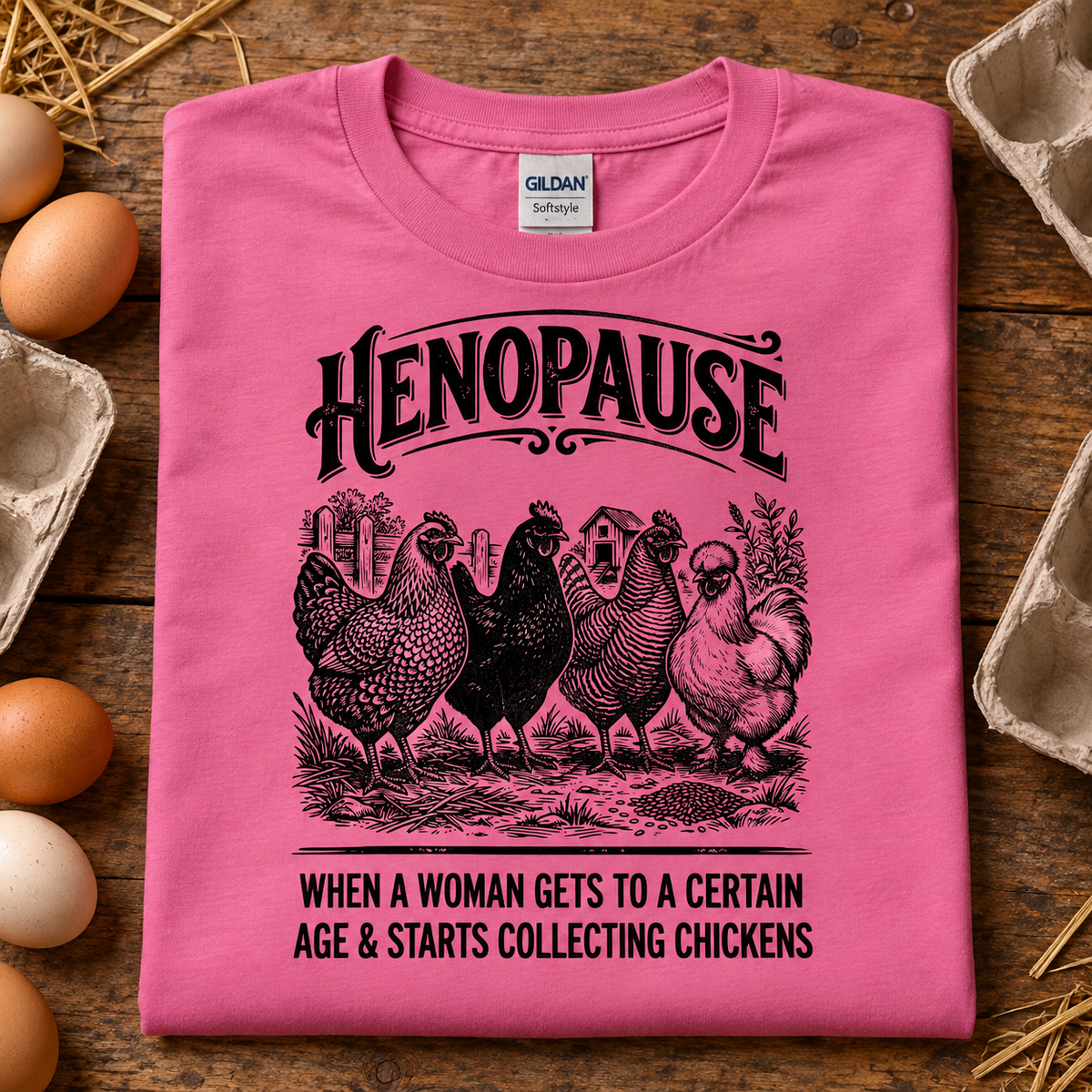 A folded azalea pink Gildan t-shirt with a humorous henopause graphic, displayed on a rustic wooden background surrounded by eggs and cardboard egg cartons. The graphic features black text reading 'HENOPAUSE' and 'WHEN A WOMAN GETS TO A CERTAIN AGE & STARTS COLLECTING CHICKENS' with an illustration of five chickens.