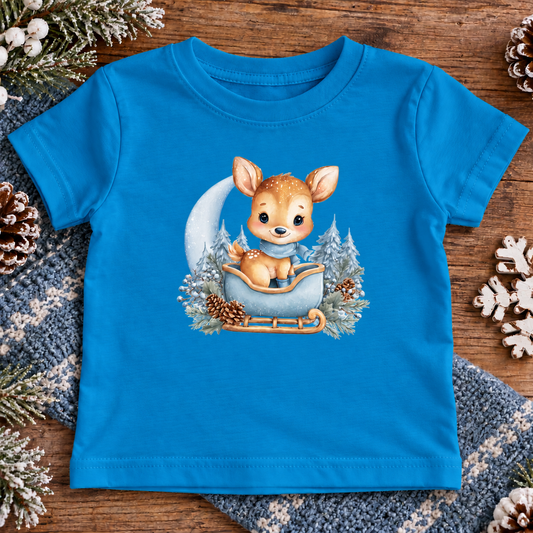 A cobalt colored toddler t-shirt with a baby deer sitting in a sleigh surrounded with trees and pinecones