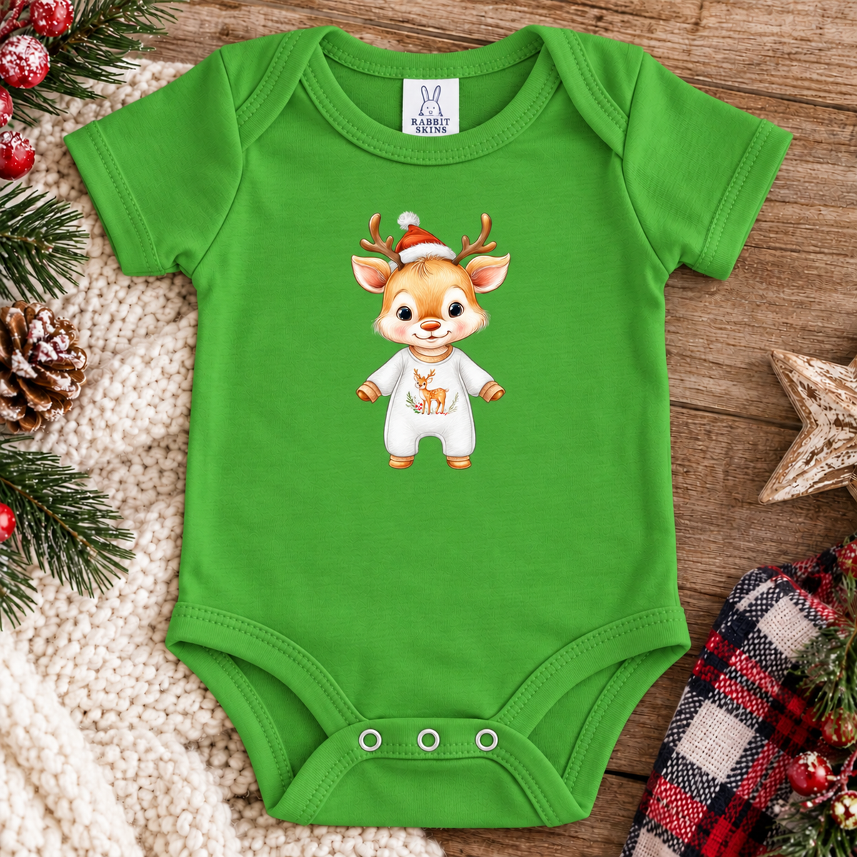 A apple green baby bodysuit with a cartoon deer graphic wearing a Santa hat and white outfit, laid flat on a wooden surface with holiday decorations around it. The neck label shows the brand 'K&H Baby'.