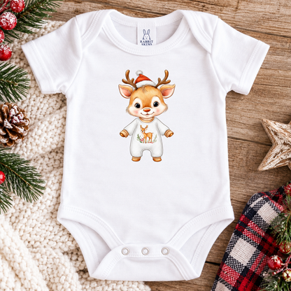 A white short-sleeve baby bodysuit laid flat on a rustic wooden surface, decorated with holiday greenery, a knit blanket, a plaid cloth, and a wooden star. The bodysuit features a cartoon deer graphic wearing a Santa hat and white outfit on the front.