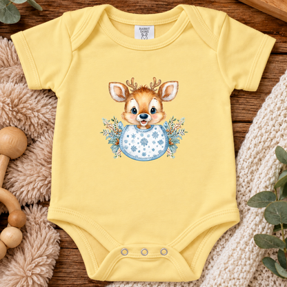 A butter yellow baby bodysuit laid flat on a wooden surface, featuring a printed illustration of a smiling deer head with antlers above a blue snowflake-patterned egg, surrounded by soft plush textures and greenery.