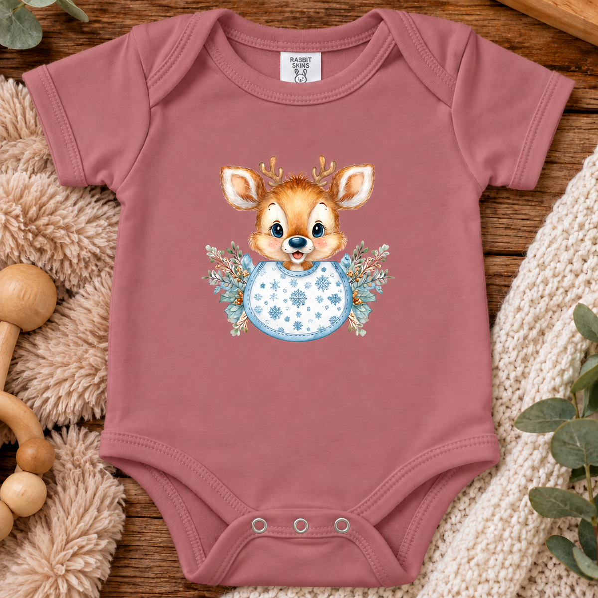 A mauvelous colored baby bodysuit laid flat on a wooden surface, featuring a cute cartoon deer graphic with a blue patterned bib and floral accents. The bodysuit is styled with cozy props including a plush toy, a knit blanket, and wooden toys.