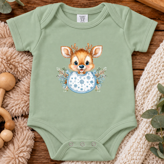 A sage green baby bodysuit laid flat on a wooden surface, featuring a cute cartoon deer graphic with a blue patterned bib and floral accents. The bodysuit is styled with a plush beige blanket, wooden toys, and a cream knit blanket.