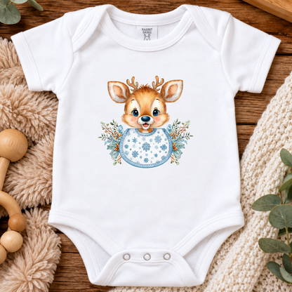 A white short-sleeve baby bodysuit laid flat on a wooden surface, featuring a watercolor-style deer graphic with antlers and a blue patterned scarf, surrounded by plush fabric, beads, and greenery.