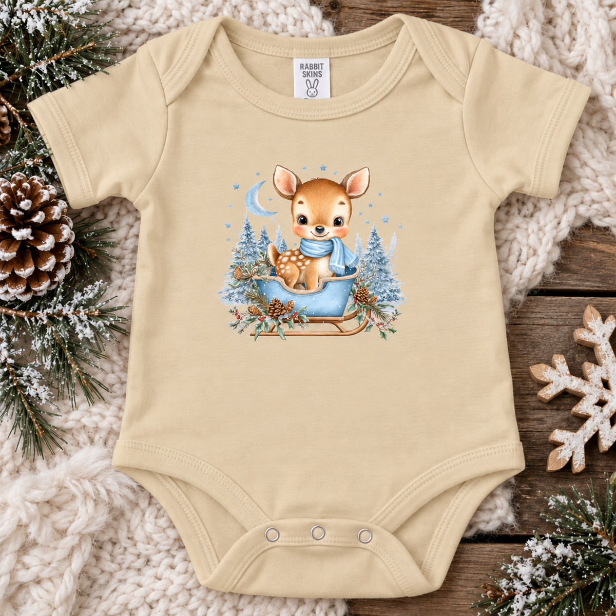 A latte baby bodysuit with a cute Christmas deer graphic, laid flat on a cozy holiday background of a knit blanket, pinecones, and wooden planks. The graphic shows a baby deer in a blue blanket and scarf sitting in a sled with pine trees.