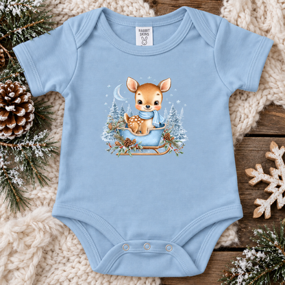 A light blue baby bodysuit laid flat on a rustic wood surface, surrounded by a chunky cream knit blanket, pine branches, a pinecone, and a decorative snowflake. The bodysuit features a central graphic of a cute brown deer sitting in a sled with a blue scarf, surrounded by snowy trees.