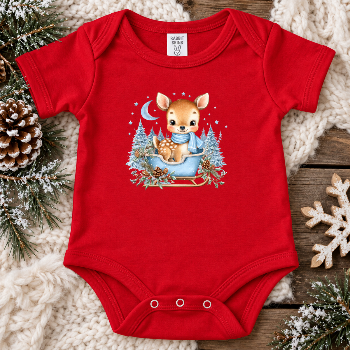 A red short-sleeve baby bodysuit laid flat on a cozy holiday-themed background. The bodysuit features a central graphic of a cute cartoon deer sitting in a blue sleigh surrounded by snowy trees and stars.