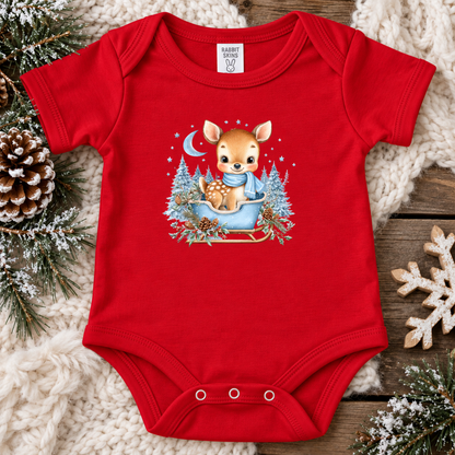 A red short-sleeve baby bodysuit laid flat on a cozy holiday-themed background. The bodysuit features a central graphic of a cute cartoon deer sitting in a blue sleigh surrounded by snowy trees and stars.