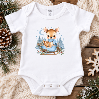A white short-sleeve baby bodysuit laid flat on a cozy knit blanket over a rustic wood surface, decorated with pine branches, a pinecone, and a snowflake. The bodysuit has a central winter illustration of a deer sitting in a blue sled with evergreen trees and a crescent moon.