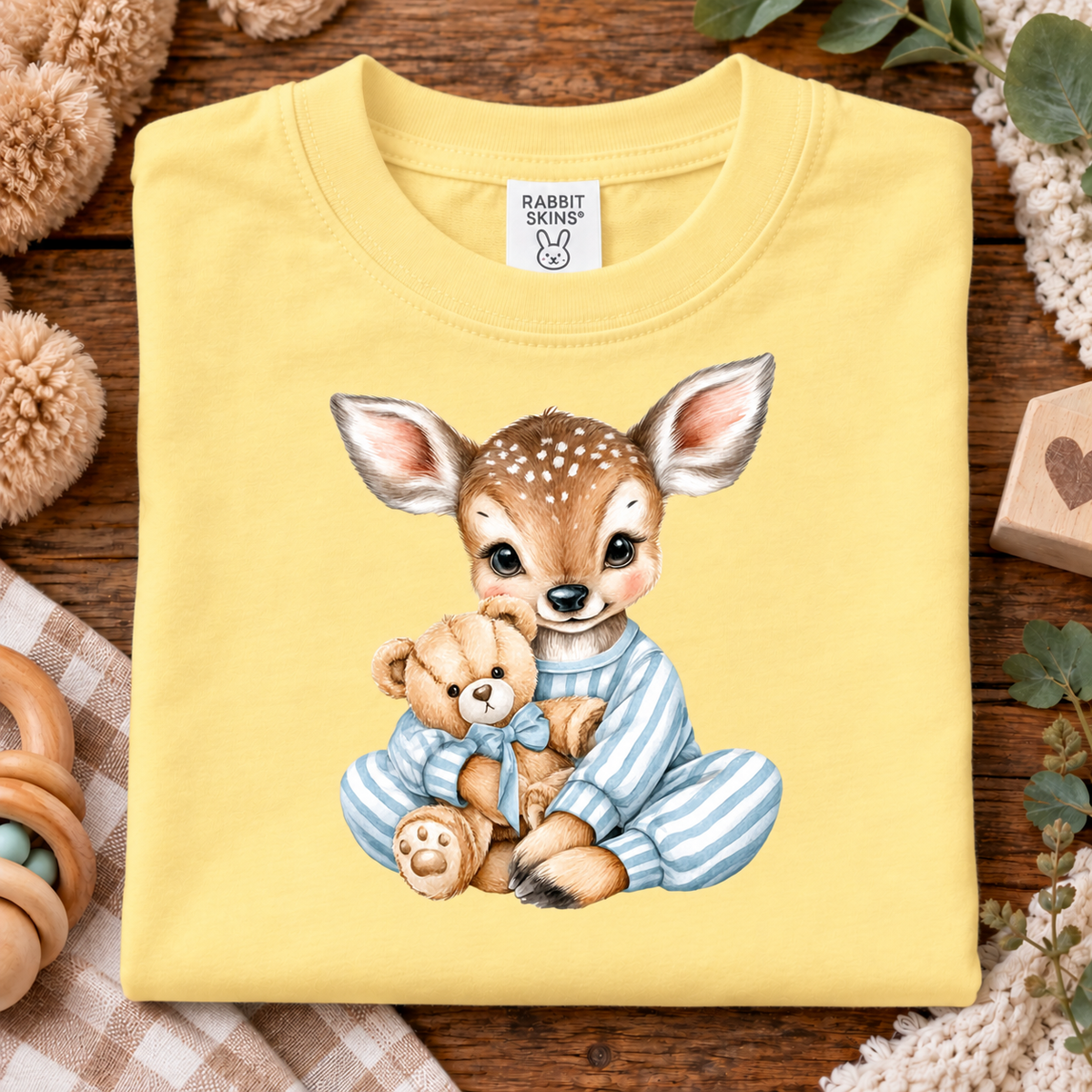 A folded butter yellow t-shirt with a cute cartoon graphic of a baby deer in blue striped pajamas hugging a teddy bear. The shirt is displayed on a rustic wooden background with baby-themed props like a wooden rattle, crochet blanket, and stuffed animal.