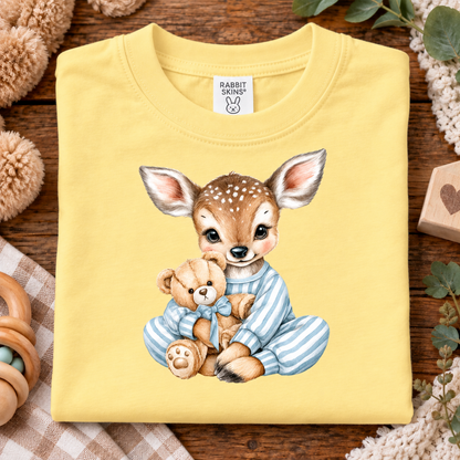 A folded butter yellow t-shirt with a cute cartoon graphic of a baby deer in blue striped pajamas hugging a teddy bear. The shirt is displayed on a rustic wooden background with baby-themed props like a wooden rattle, crochet blanket, and stuffed animal.