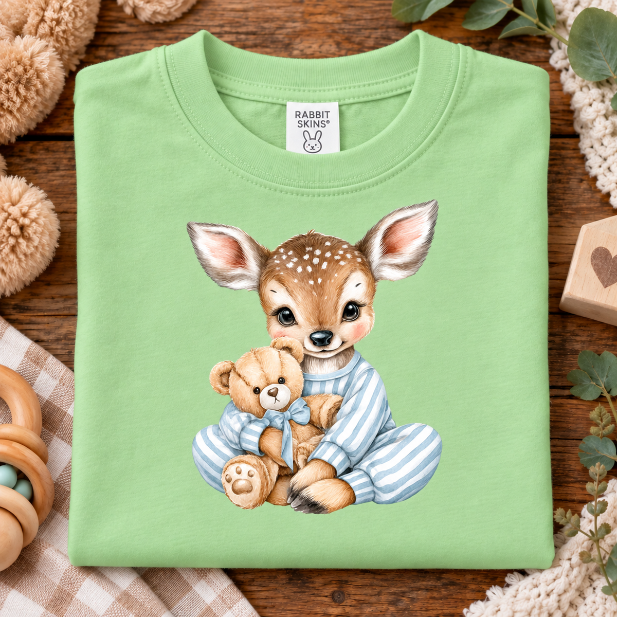 A folded keylime t-shirt with a cute watercolor illustration of a baby deer in blue striped pajamas hugging a teddy bear, displayed on a rustic wooden background with cozy baby accessories.
