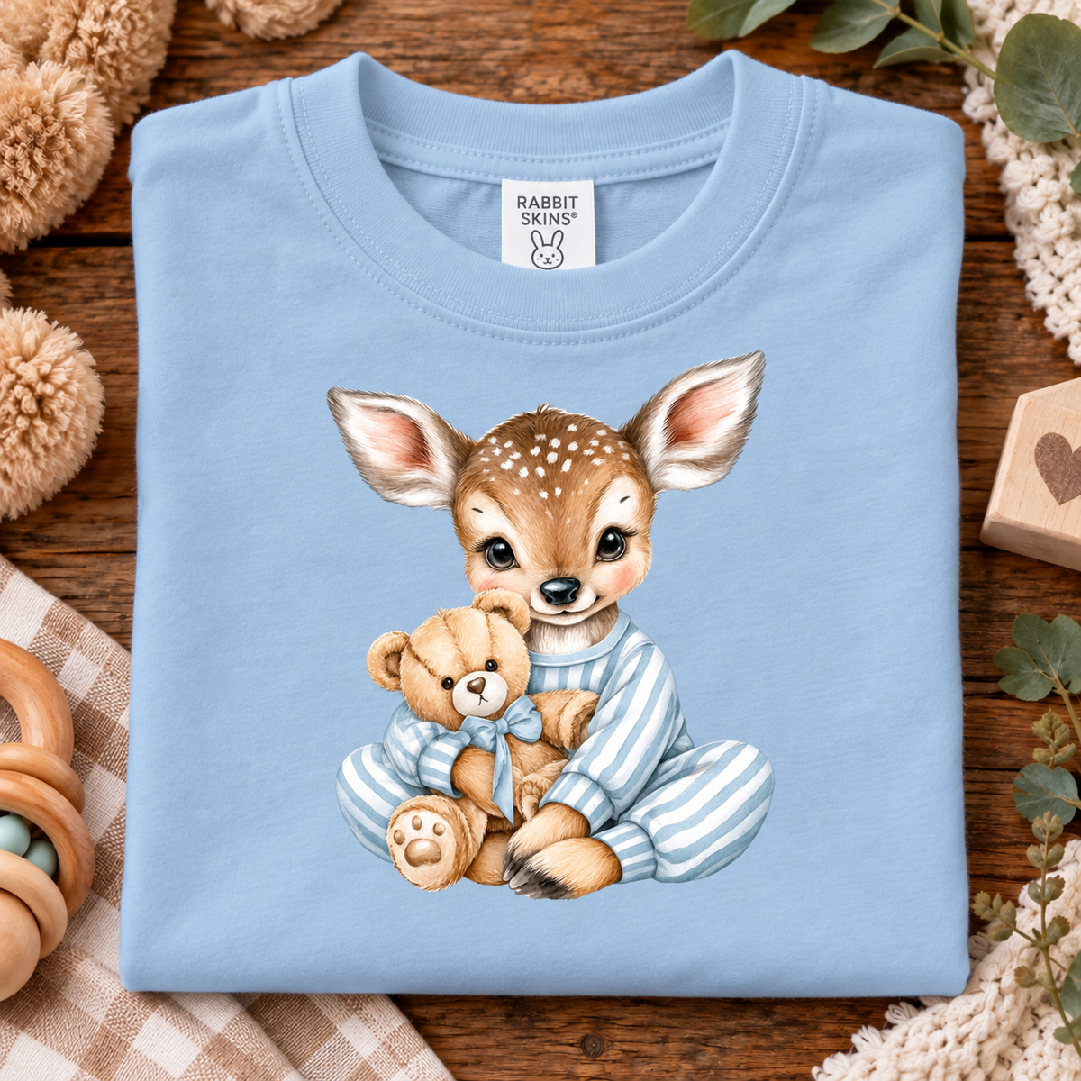 A light blue baby t-shirt with a watercolor graphic of a fawn in striped pajamas holding a teddy bear, laid flat on a rustic wooden background with baby-themed props.