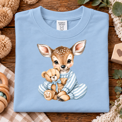 A light blue baby t-shirt with a watercolor graphic of a fawn in striped pajamas holding a teddy bear, laid flat on a rustic wooden background with baby-themed props.