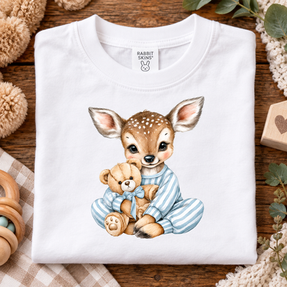 A folded white short-sleeve baby T-shirt with a cute watercolor-style deer fawn graphic holding a teddy bear, styled on a wooden surface with soft baby props.