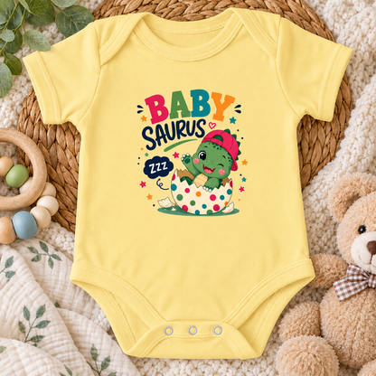 A butter yellow short-sleeve baby bodysuit laid flat on a woven mat, surrounded by a wooden ring toy, green leaves, a teddy bear, and a soft blanket. The bodysuit has a colorful graphic that reads “BABY SAURUS” with a cartoon green dinosaur sitting in a polka-dot teapot and a “zzz” speech bubble.