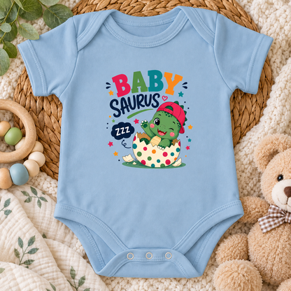 A light blue baby bodysuit laid flat on a woven mat, featuring a colorful 'BABY SAURUS' graphic with a cartoon dinosaur hatching from an egg. The bodysuit is styled with a wooden ring toy, a plush teddy bear, and a soft cream blanket.