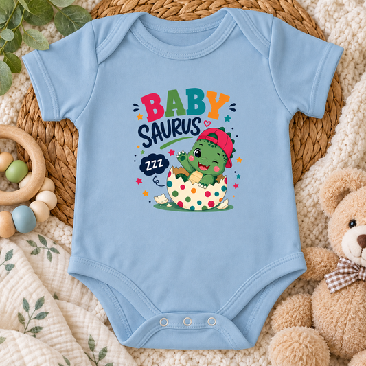 A light blue baby bodysuit laid flat on a woven mat, featuring a colorful 'BABY SAURUS' graphic with a cartoon dinosaur hatching from an egg. The bodysuit is styled with a wooden ring toy, a plush teddy bear, and a soft cream blanket.