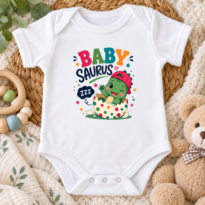 A white short-sleeve baby bodysuit laid flat on a woven mat, surrounded by a wooden ring toy, green leaves, a teddy bear, and a cream blanket. The bodysuit features a colorful graphic that reads 'BABY SAURUS' above a cartoon green dinosaur sitting in a polka-dot bassinet.