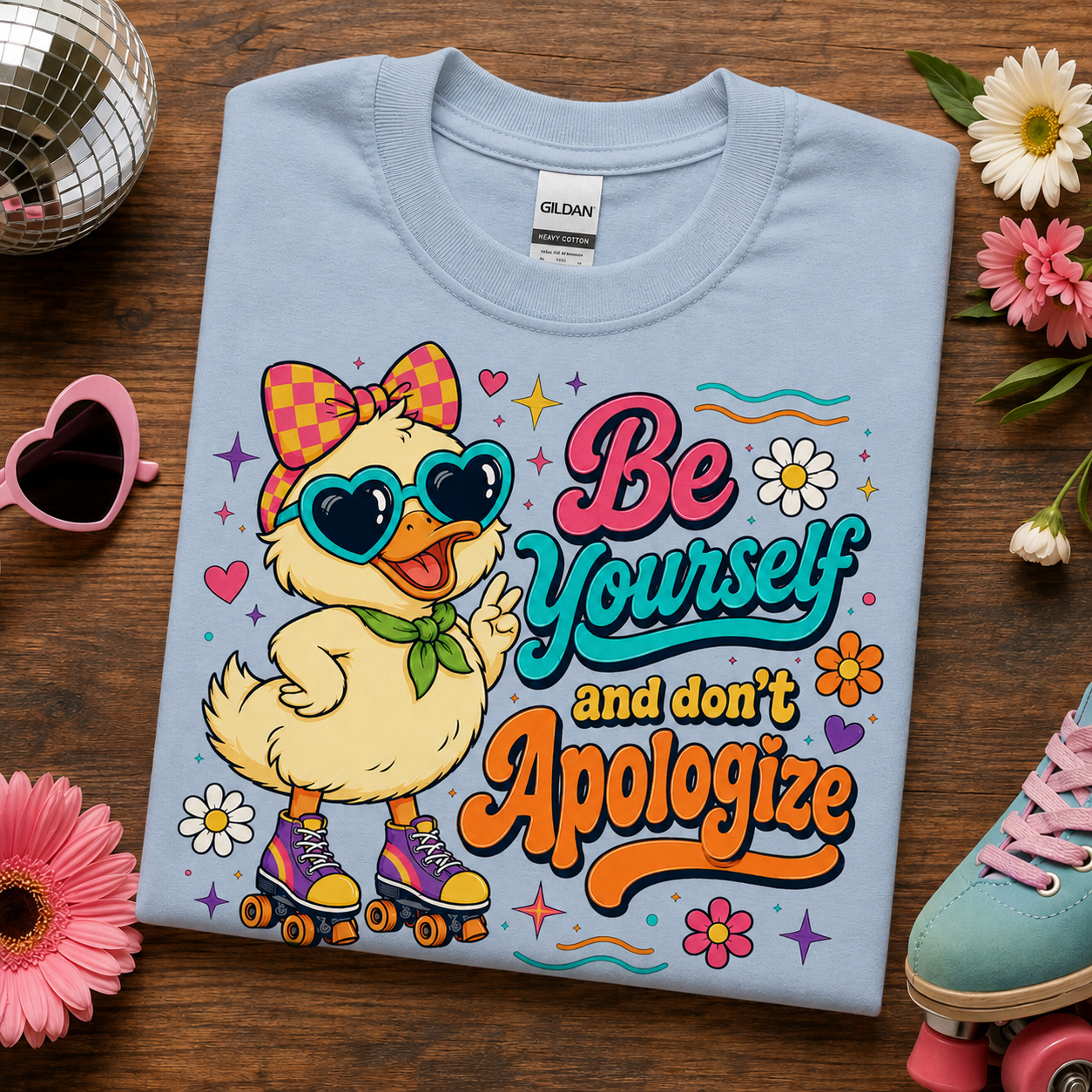A folded light blue Gildan t-shirt is displayed on a dark wood background, surrounded by decorative lifestyle props including a disco ball, pink sunglasses, flowers, and a pastel shoe. The shirt features a colorful graphic of a yellow duck character wearing heart-shaped sunglasses and roller skates, with the text 'Be Yourself and don't Apologize'.