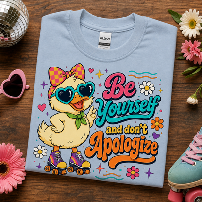 A folded light blue Gildan t-shirt is displayed on a dark wood background, surrounded by decorative lifestyle props including a disco ball, pink sunglasses, flowers, and a pastel shoe. The shirt features a colorful graphic of a yellow duck character wearing heart-shaped sunglasses and roller skates, with the text 'Be Yourself and don't Apologize'.