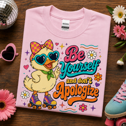 A folded light pink Gildan t-shirt with a colorful graphic of a duck character on roller skates and the text 'Be Yourself and don't Apologize', styled on a dark wood background with flowers, a disco ball, pink heart sunglasses, and a blue shoe.