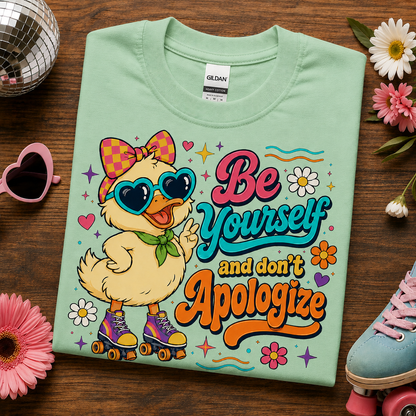 A mint green Gildan t-shirt laid flat on a dark wooden surface, featuring a colorful graphic of a yellow duck character wearing heart-shaped sunglasses and roller skates, with the text 'Be Yourself and don't Apologize'. The shirt is styled with pink and white daisies, a pink heart-shaped sunglasses, a disco ball, and a blue roller skate.
