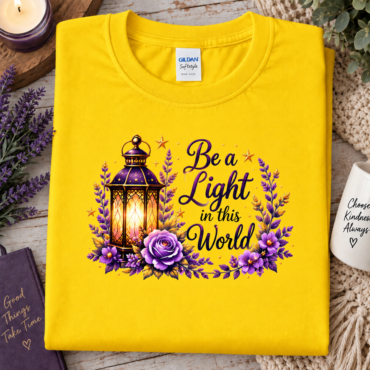 A folded daisy yellow Gildan Softstyle T-shirt with a graphic print of a glowing lantern, purple flowers, and the text 'Be a Light in this World,' styled on a rustic wooden surface with candles, lavender, and a mug nearby.