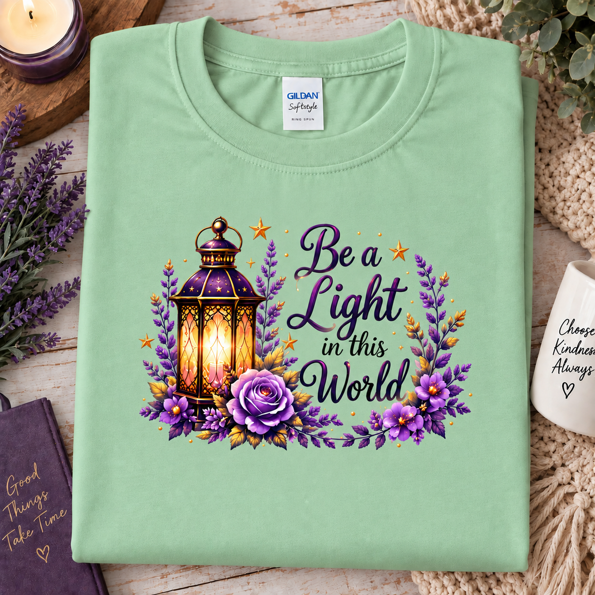 A folded mint green Gildan t-shirt is displayed on a rustic wooden surface surrounded by decorative props. The front of the shirt features a graphic print with a glowing lantern, purple flowers, and the text 'Be a Light in this World'.