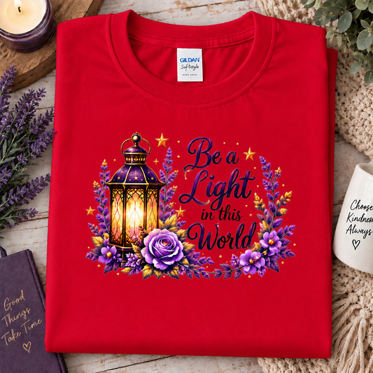 A folded red Gildan t-shirt is displayed on a rustic wooden surface surrounded by decorative props. The front of the shirt features a graphic print with a glowing lantern, purple flowers, and the text 'Be a Light in this World'.