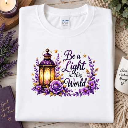 A folded white Gildan Softstyle t-shirt with a graphic print of a glowing lantern, purple flowers, and the text 'Be a Light in this World'. The shirt is styled in a cozy flat-lay setting with a candle, a notebook, and a mug on a wooden surface.