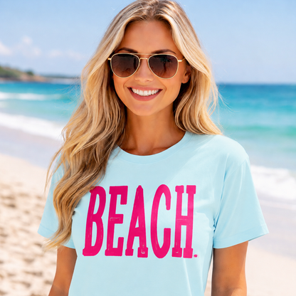 Woman wearing a chambray colored t-shirt with 'BEACH' in pink on a beach.