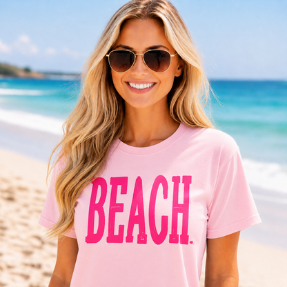 Woman wearing a blossom colored t-shirt with 'BEACH' in pink on a beach.