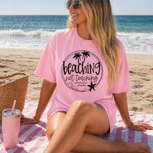 Woman in pink 'Beaching not teaching' shirt sitting on a beach with ocean view.