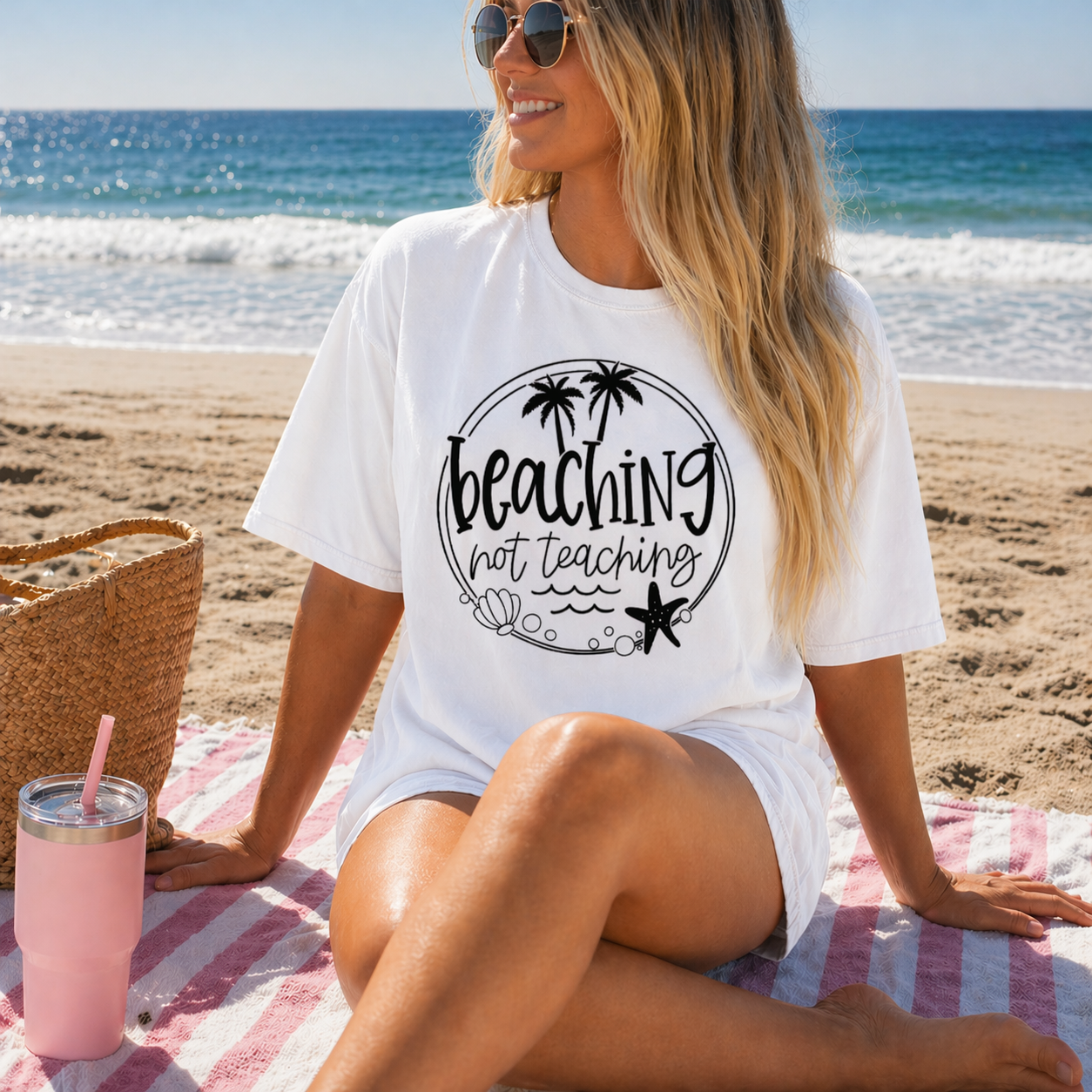 Woman sitting on a beach with a white shirt that says 'Beaching not teaching'.
