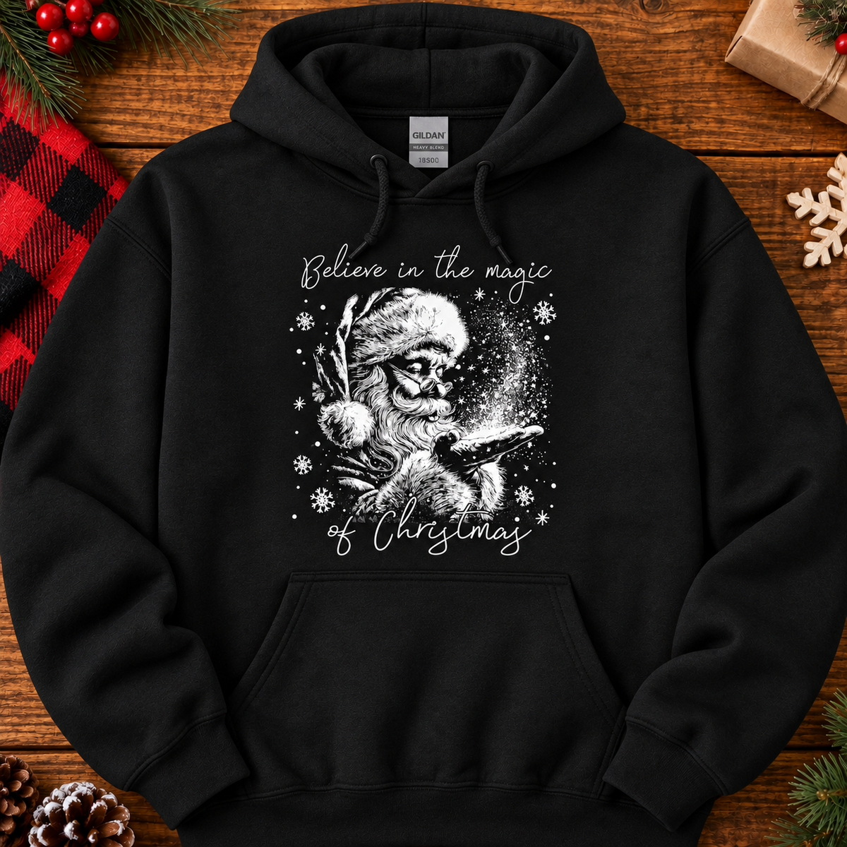 A black pullover hoodie laid flat on a rustic wooden surface, featuring a white graphic of a Santa skull holding a glowing Christmas tree, with the text 'Believe in the magic of Christmas.' The hoodie is styled with holiday props including pine branches, a red plaid blanket, a gift box, and a snowflake ornament.