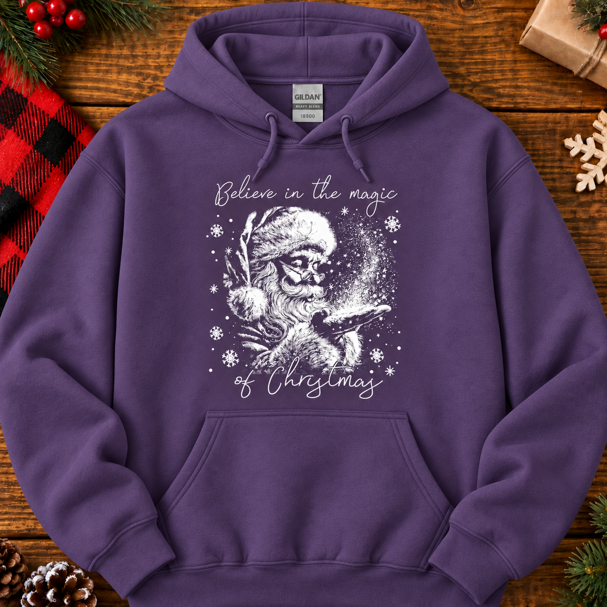 A purple pullover hoodie laid flat on a rustic wooden surface, decorated with holiday props. The hoodie features a white graphic of a Santa skeleton holding a glowing Christmas tree, with the text 'Believe in the magic of Christmas' written around the design.
