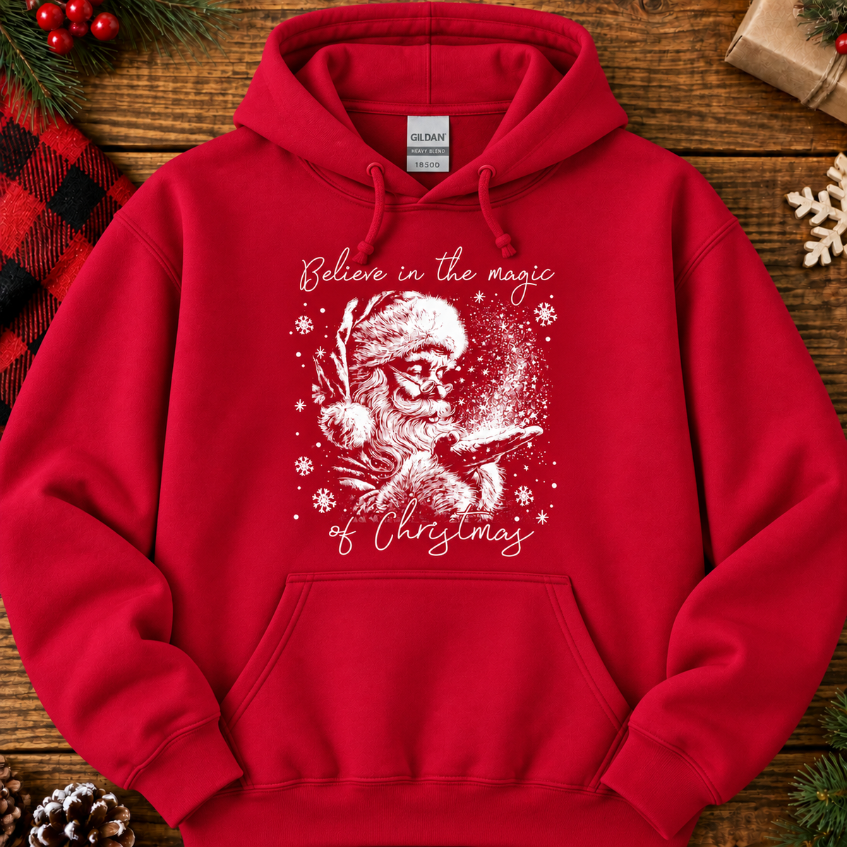 A red pullover hoodie laid flat on a rustic wooden surface with holiday decorations around it. The hoodie has a white graphic of Santa with a sleigh and snowflakes, with the text 'Believe in the magic of Christmas' printed on the front.
