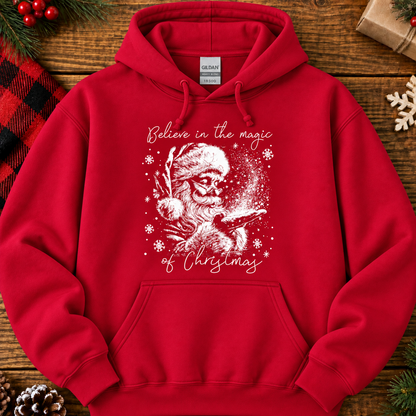 A red pullover hoodie laid flat on a rustic wooden surface with holiday decorations around it. The hoodie has a white graphic of Santa with a sleigh and snowflakes, with the text 'Believe in the magic of Christmas' printed on the front.