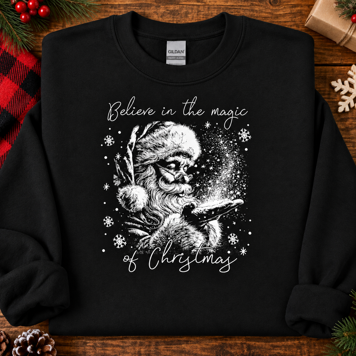 A black long-sleeve crewneck sweatshirt with a white graphic of Santa Claus and a bird, surrounded by the text 'Believe in the magic of Christmas'. The sweatshirt is displayed flat on a rustic wooden background with holiday decorations.