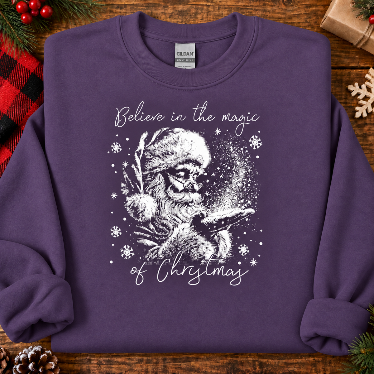 A folded purple crewneck sweatshirt with a white graphic of Santa Claus and the text 'Believe in the magic of Christmas', displayed on a rustic wooden background with holiday decorations.