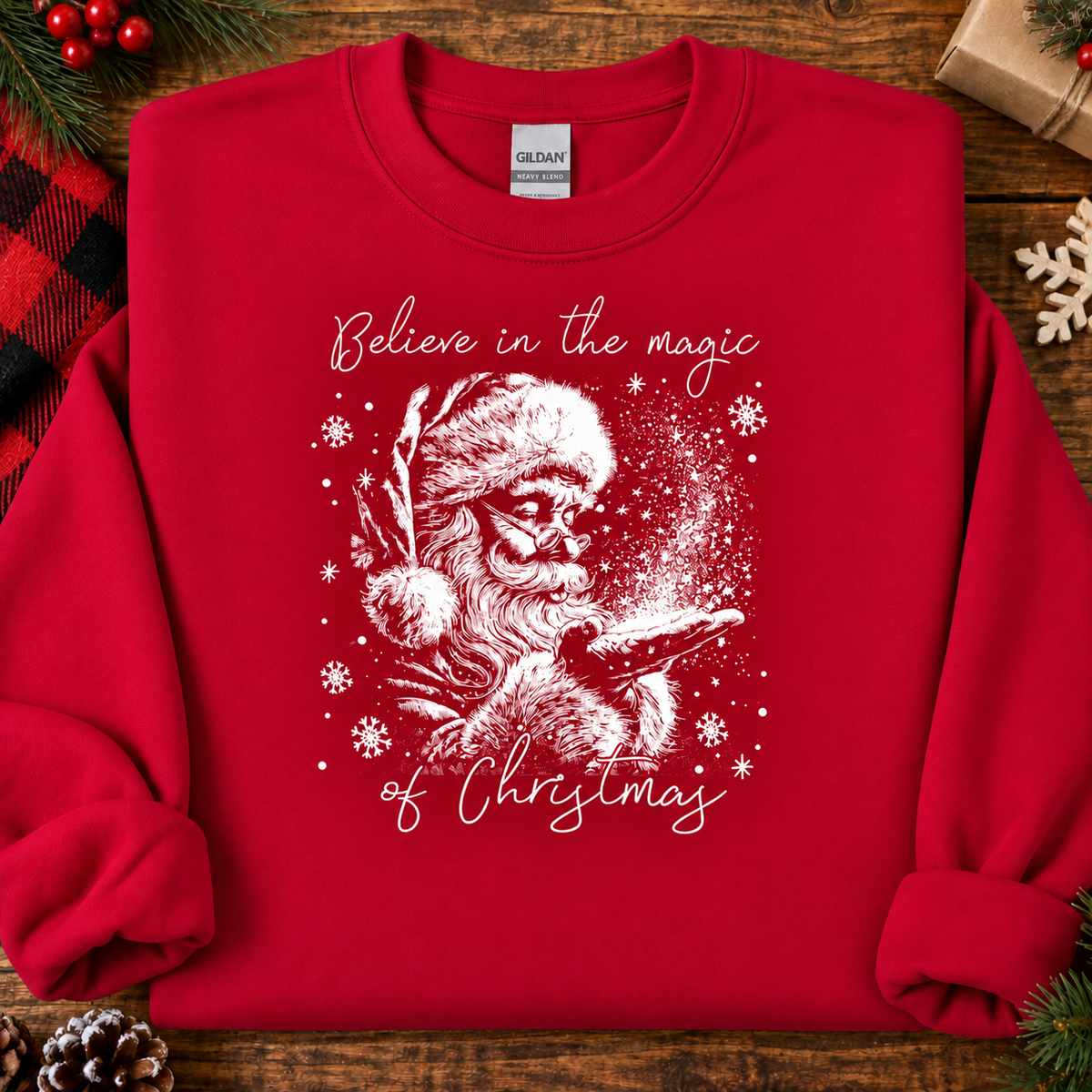 A red crewneck sweatshirt with a white graphic of Santa Claus and the text 'Believe in the magic of Christmas' laid flat on a rustic wooden surface with holiday decorations.