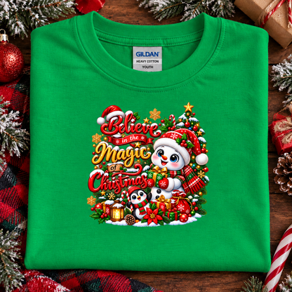 A folded green Gildan T-shirt with a colorful Christmas graphic reading 'Believe in the Magic of Christmas' and featuring cartoon snowman, penguin, ornaments, and holiday decorations, displayed on a rustic wood surface with festive props.