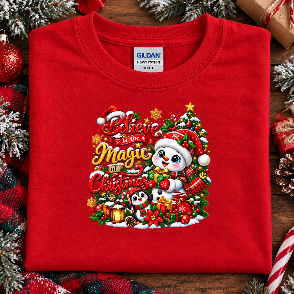 A folded red Gildan youth t-shirt with a colorful Christmas graphic on the front, displayed on a rustic wooden background decorated with pine branches, ornaments, and wrapped gifts.