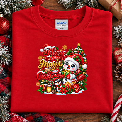 A folded red Gildan youth t-shirt with a colorful Christmas graphic on the front, displayed on a rustic wooden background decorated with pine branches, ornaments, and wrapped gifts.