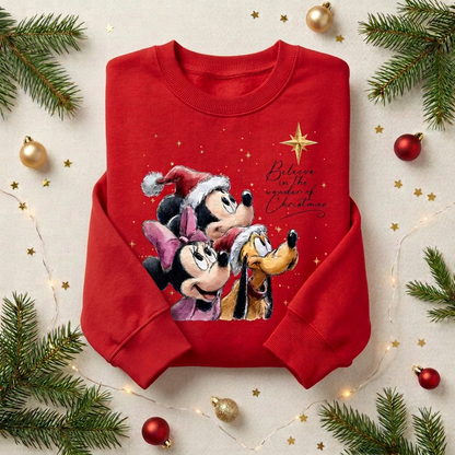 A red crewneck sweatshirt featuring a festive Christmas graphic of Mickey Mouse, Minnie Mouse, and Pluto wearing Santa hats, with the text 'Believe in the spirit of Christmas' and a gold star. The sweatshirt is displayed in a flat-lay arrangement with pine branches, gold ornaments, and fairy lights on a light beige background.