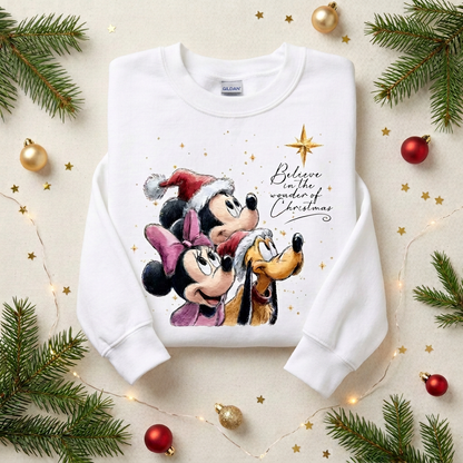 A white long-sleeve sweatshirt laid flat on a light beige surface, decorated with pine branches, gold ornaments, and fairy lights. The sweatshirt features a colorful graphic of Mickey Mouse, Minnie Mouse, Pluto, and Santa with the text 'Believe in the wonder of Christmas.